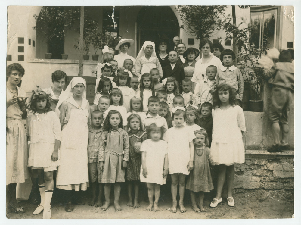 Photographie d'un «premier groupe d'enfants orphelins après les turbulences de la guerre», encadrés par des infirmières et autres adultes – avec légende manuscrite et tampons de la Croix Blanche polonaise au verso